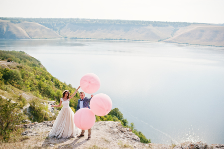 Gorgeous bride and groom having great time standing on the precipice with a view of a lake with balloons in their hands on their wedding day.の写真素材