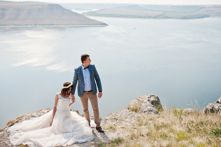 Gorgeous bride sitting on the stone while her husband stands next to her on the cliff with lake in the background.の写真素材
