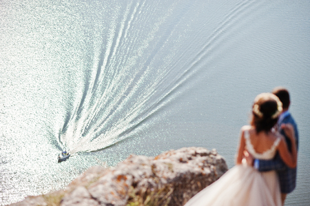 Awesome wedding couple admiring view of lake and boat sailing from the rocky cliff.の写真素材