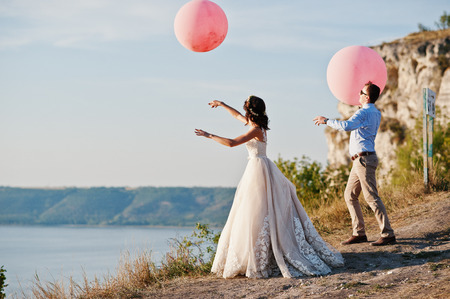 Gorgeous bride and groom having great time standing on the precipice with a view of a lake with balloons in their hands on their wedding day.の写真素材
