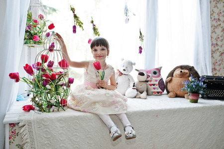 Cute little girl wearing amazing dress sitting on the windowsill next to the flowers with a tupil in her hand.の写真素材