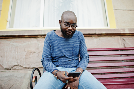 Black african american guy sitting on the purple wooden bench on the street wearing casual clothes.の写真素材