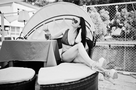 Portrait of a stunning girl in red bikini swimsuit and sunglasses enjoying her time by sitting on the sofa on the quay by the lake. Black and white photo.の写真素材