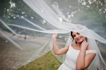 Portrait of a gorgeous bride posing with her long veil in the park on the wedding day.