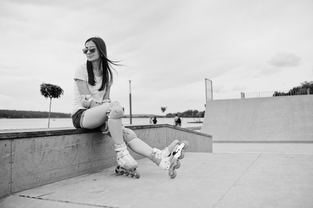 Portrait of an attractive young woman in shorts, t-shirt, sunglasses and rollerblades sitting on the concrete bench in the outdoor roller skating rink. Black and white photo.の写真素材