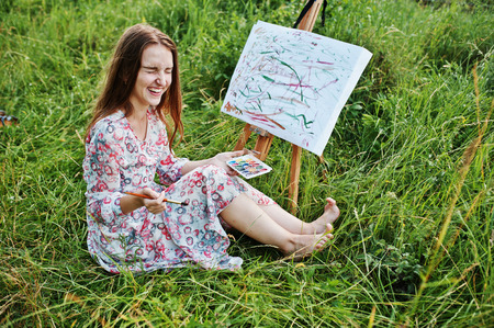 Portrait of a gorgeous happy young woman in beautiful dress sitting on the grass and painting on paper with watercolors.の写真素材