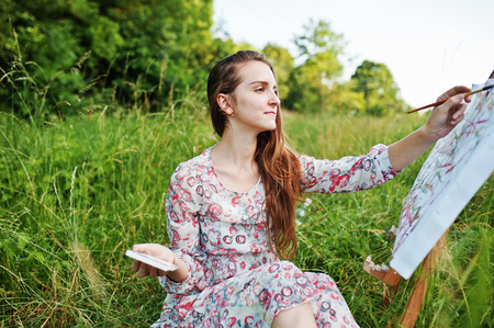 Portrait of a gorgeous happy young woman in beautiful dress sitting on the grass and painting on paper with watercolors.の写真素材