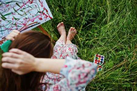 Portrait of a gorgeous happy young woman in beautiful dress sitting on the grass and painting on paper with watercolors.の写真素材