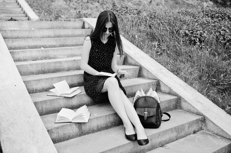 Portrait of a beautiful woman in black polka dot dress and sunglasses sitting on the stairs with books and backpack in the park. Black and white photo.の写真素材