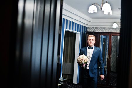 Portrait of a handsome groom posing with bridal bouquet in his cool room on the wedding day.の写真素材