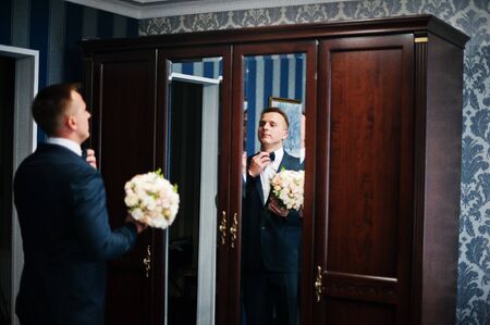 Portrait of a handsome groom posing with bridal bouquet in his cool room on the wedding day.の写真素材