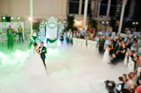 Beautiful wedding couple dancing their first dance in the huge hall with heavy smoke, different...