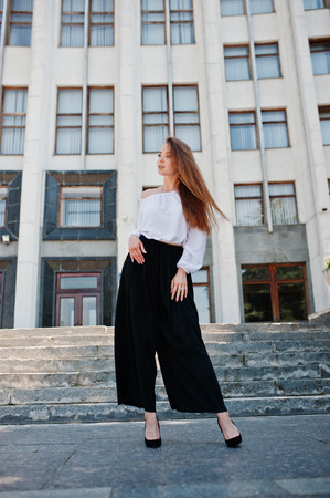 Portrait of a fabulous young successful woman in white blouse and broad black pants posing on the stairs with a huge white building on the background.の写真素材