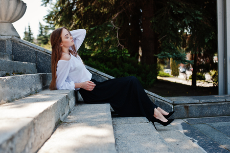 Good-looking young woman in white blouse, wide black pants and black classic high heels sitting on stairs and posing.の写真素材