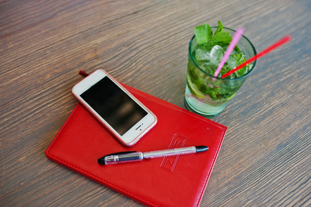 Close-up photo of a red notebook, black pen, smartphone and a cocktail on a table in a cafe.の写真素材