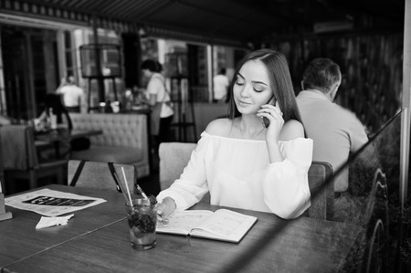 Portrait of a gorgeous young businesswoman talking on the phone and writing something down in her red notebook while sitting in a cafe. Black and white photo.の写真素材