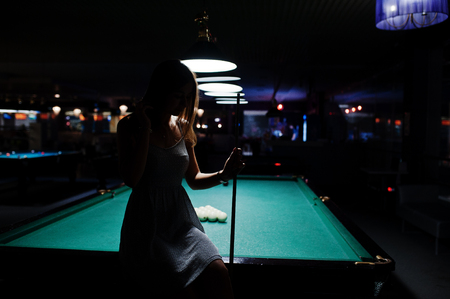 Portrait of an attractive young woman in dress sitting on the pool table and holding a cue.の写真素材