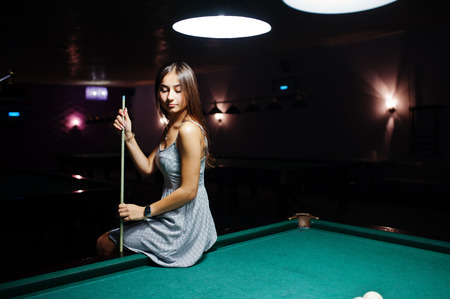 Portrait of an attractive young woman in dress sitting on the pool table and holding a cue.の写真素材