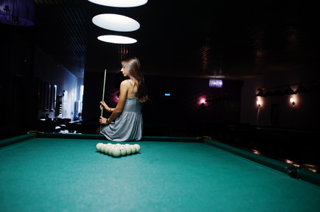 Portrait of an attractive young woman in dress sitting on the pool table and holding a cue.