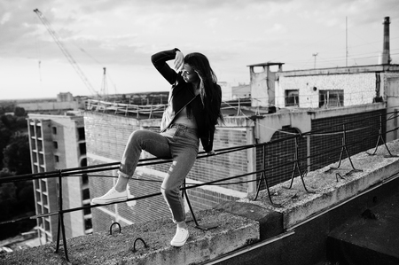 Portrait of a gorgeous young woman in black leather jacket, jeans and sneakers sitting on handrails on the roof with picturesque view of a park. Black and white photo.の写真素材