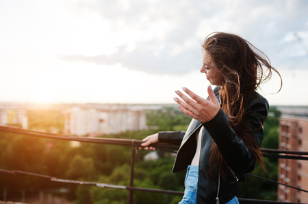 Portrait of an attractive young woman in black leather jacket, jeans and sneakers standing by the handrails on the rooftop.の写真素材