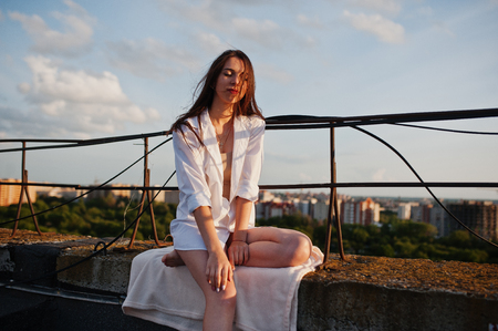 Portrait of a fabulous young woman in white male shirt sitting on the edge of the building roof with a fantastic view of a city in a background.の写真素材