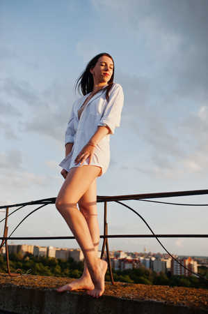 Portrait of an outstanding woman in white male shirt posing on top of the building with charming scenery on the background.の写真素材
