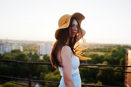 Portrait of a pretty young woman in white t-shirt and blue skirt posing on the rooftop with her orange hat at the sunset.の写真素材