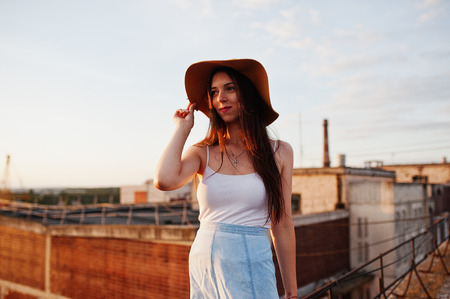 Portrait of a pretty young woman in white t-shirt and blue skirt posing on the rooftop with her orange hat at the sunset.の写真素材