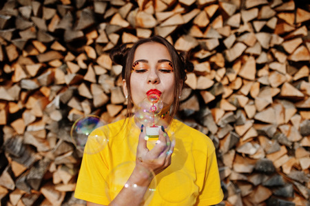 Young funny girl with bright make-up, wear on yellow shirt blown soap bubbles against wooden background.の写真素材