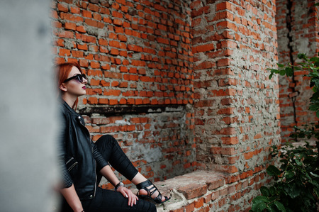 Red haired stylish girl in sunglasses wear in black, against abadoned place with brick walls.の写真素材