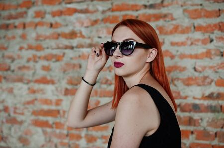 Red haired stylish girl in sunglasses wear in black, against abadoned place with brick walls.の写真素材