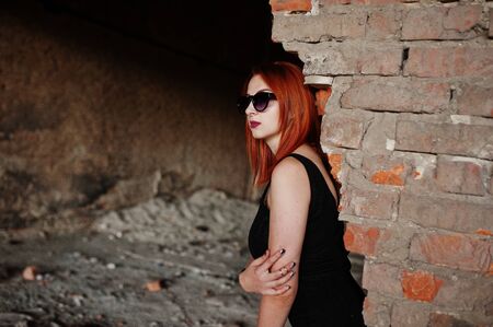Red haired stylish girl in sunglasses wear in black, against abadoned place with brick walls.の写真素材