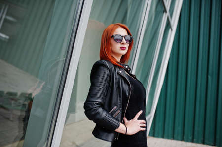 Red haired stylish girl in sunglasses wear in black, against large windows. Street fashion portrait.の写真素材