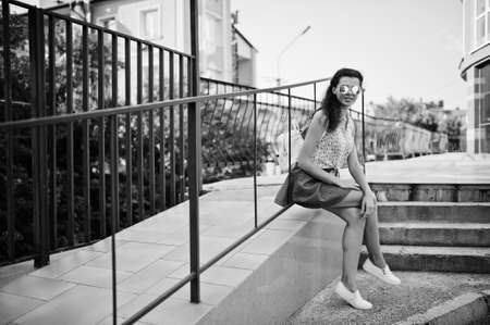 Curly stylish girl wear on blue jeans skirt, blouse sitting on bench. Portrait on streets of city.の写真素材