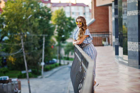 Portrait of a perfect young woman wearing striped overall and yellow sunglasses poses on a balcony of a modern building in a town.の写真素材