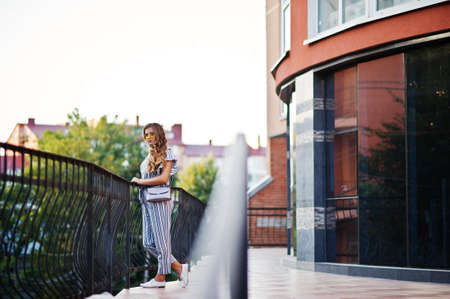 Portrait of a perfect young woman wearing striped overall and yellow sunglasses poses with her handbag on a balcony of a modern building in a town.の写真素材