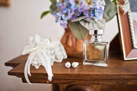 Bridal perfume, white lace garter and elegant wedding earrings on the table with flowers in the background.の写真素材