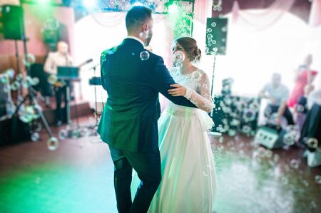 Beautiful wedding couple performing their first dance in the restaurant with different lights and bubbles and guests on the background.の写真素材