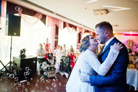 Beautiful wedding couple performing their first dance and kissing in the restaurant with different lights and bubbles and guests on the background.の写真素材