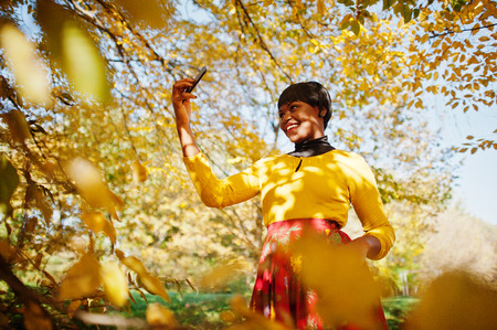 African american girl at yellow and red dress at golden autumn fall park making selfie on phone.の写真素材