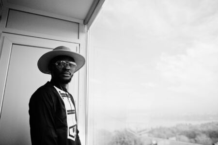 Rich african man looking at panoramic window at his penthouse. Portrait of successful black man in hat indoor.の写真素材