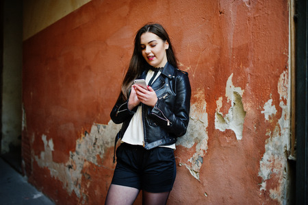 Stylish brunette girl wear on leather jacket and shorts with mobile phone at hands against orange wall.の写真素材