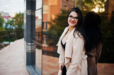Black hair sexy woman in glasses and coat posed against building with modern windows.の写真素材