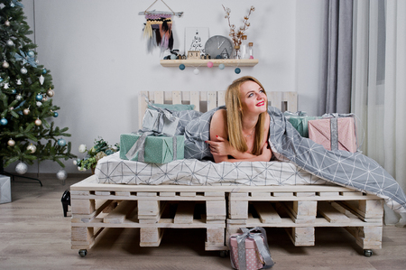 Cute blonde girl on black bra lying on bed with gifts boxes against new year tree at studio. Happy holidays.の写真素材