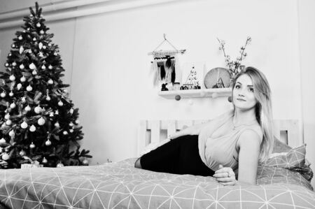Blonde girl in dress posed on bed near new year tree with christmas gifts decor in white room.の写真素材