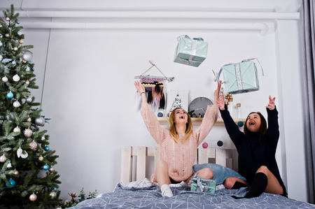 Two fun beautiful girls friends wear in warm sweater and leg warmers (gaiters) with gifts boxes on bed against new year tree with christmas decoration.の写真素材