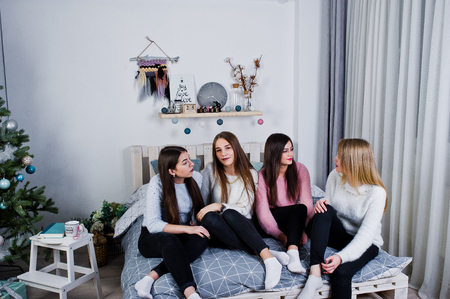 Four cute friends girls wear on warm sweaters and black pants on bed at new year decorated room on studio.の写真素材