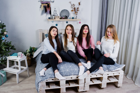Four cute friends girls wear on warm sweaters and black pants on bed at new year decorated room on studio.の写真素材