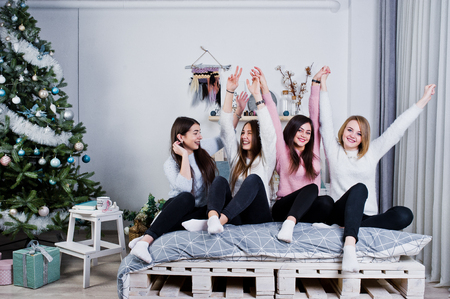 Four cute friends girls wear on warm sweaters and black pants on bed at new year decorated room on studio.の写真素材